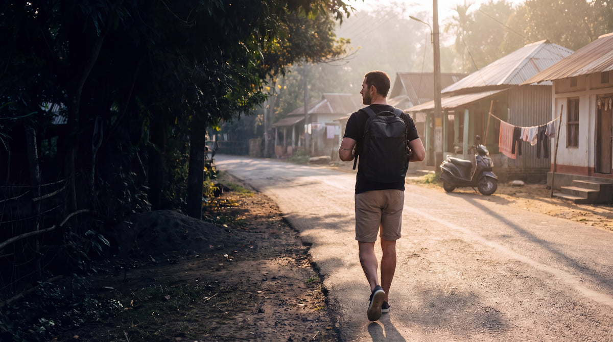 A man in a black HercShirt with a compact travel backpack walking along a quiet village road, seen from behind